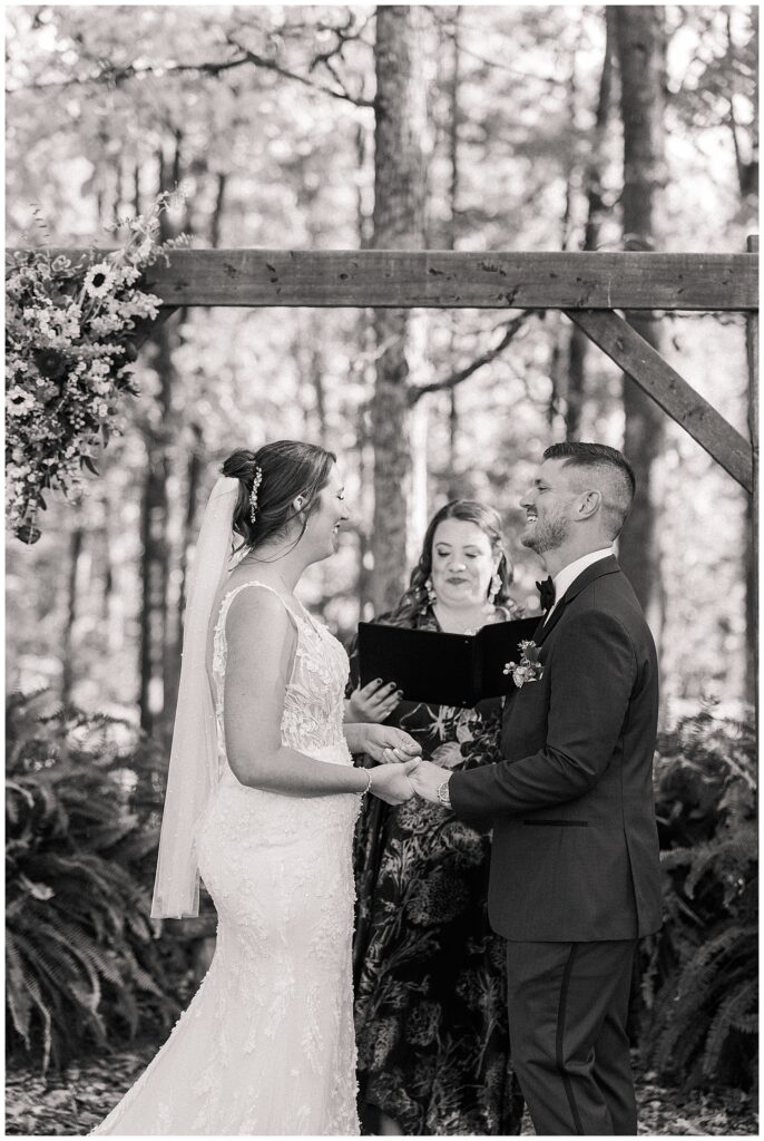 Bride and groom exchange heartfelt vows under a wooden arch with their female officiant during the intimate forest ceremony.