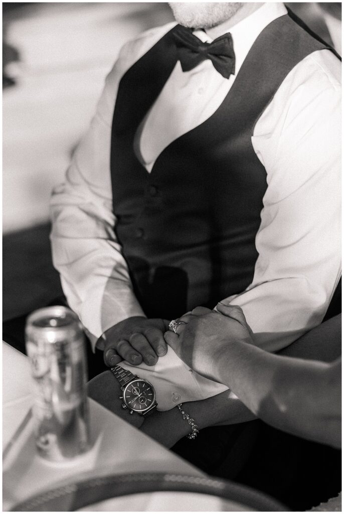 Close-up black and white photo of the bride and groom holding hands across the table, highlighting their rings and connection.