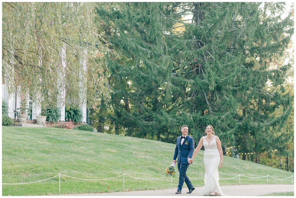 Emily and Tommy walk side-by-side on the grassy lawn in front of tall evergreens at Athena Estate’s scenic property.