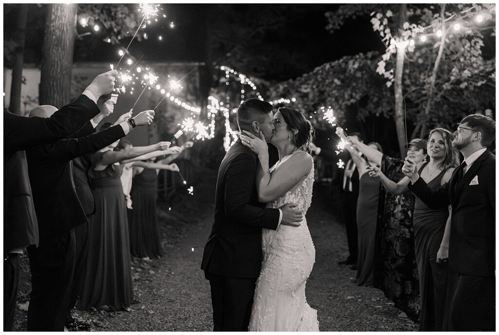 Bride and groom share a romantic kiss surrounded by guests holding sparklers during their nighttime send-off.