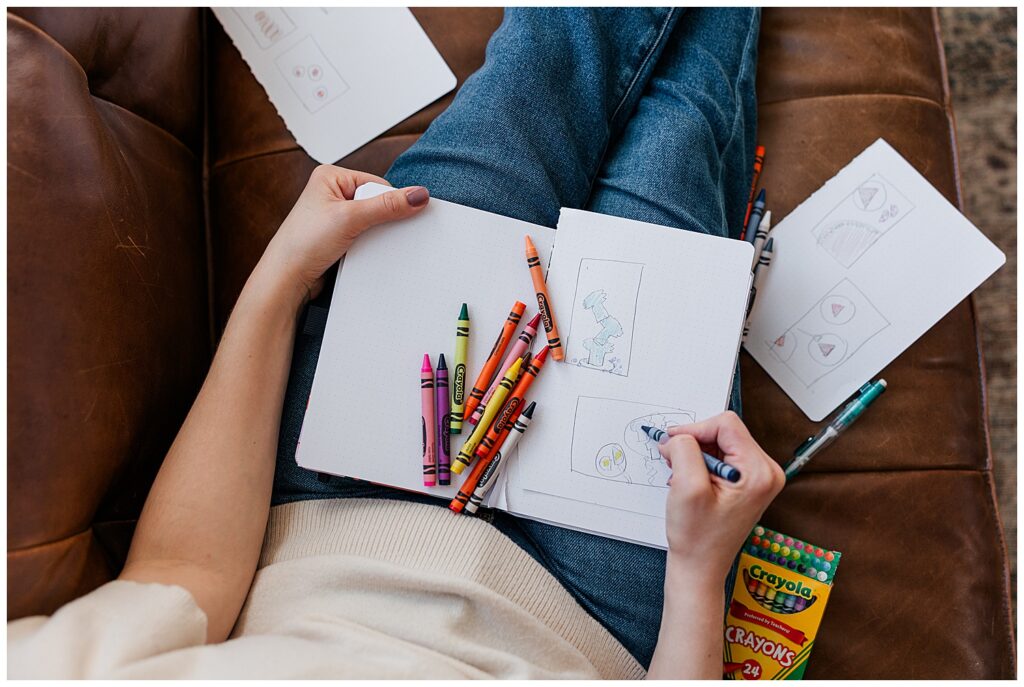 Overhead view of a person drawing with colorful crayons in a sketchbook while sitting cross-legged on a couch, highlighting creativity and relaxation.