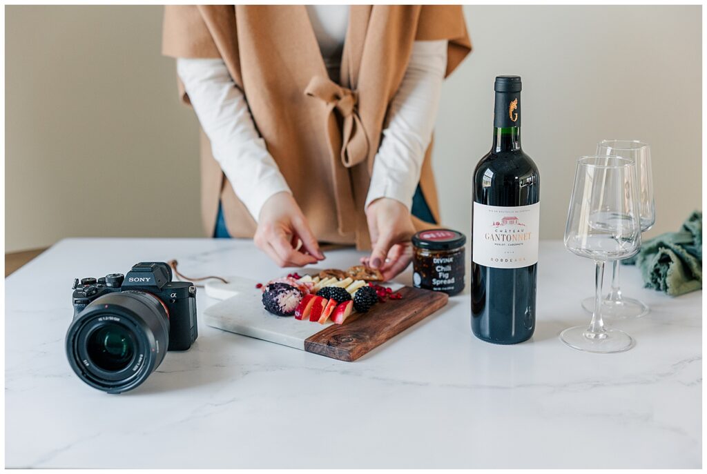 Flat lay style brand photo of Ashleigh arranging a charcuterie board with fruit, jam, and wine on a marble counter, showcasing a cozy lifestyle brand aesthetic.