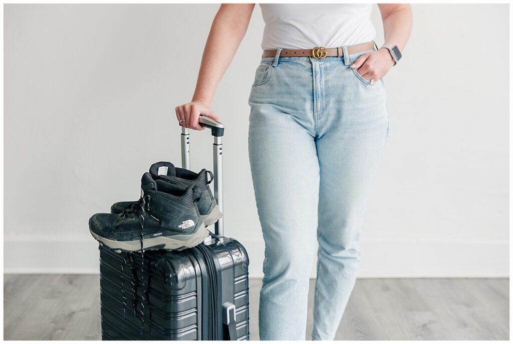 Close-up brand photo of a woman holding a suitcase and black heels, ready to travel in casual jeans and a belt.