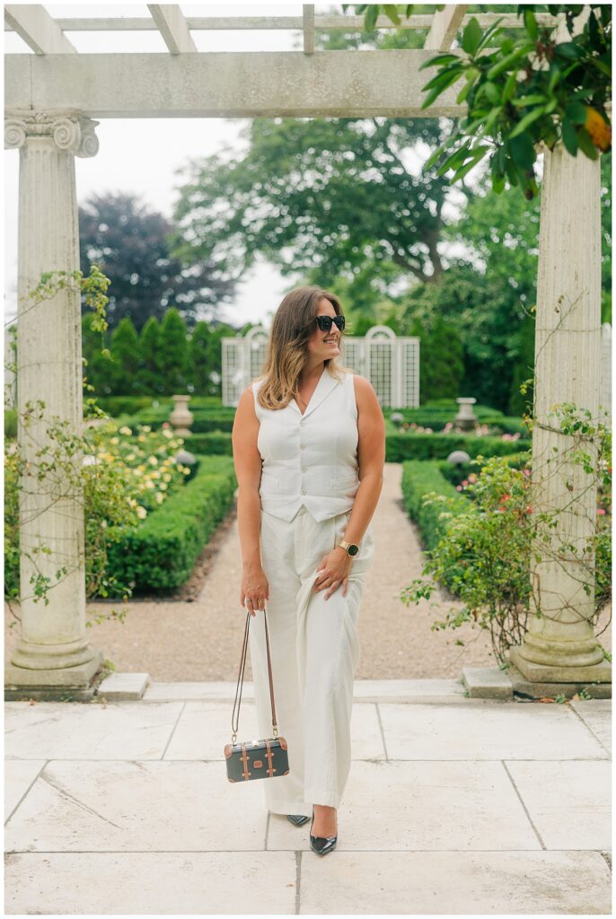 Luxury travel branding photo of a woman walking through a rose garden in a white outfit and sunglasses at a Newport estate.