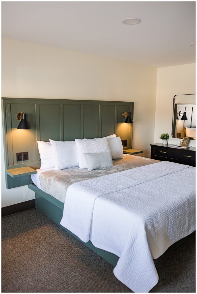 Interior photo of a modern hotel room featuring a king-size bed with white linens, sage green headboard, and warm bedside lighting, perfect for Airbnb or travel brand photography.