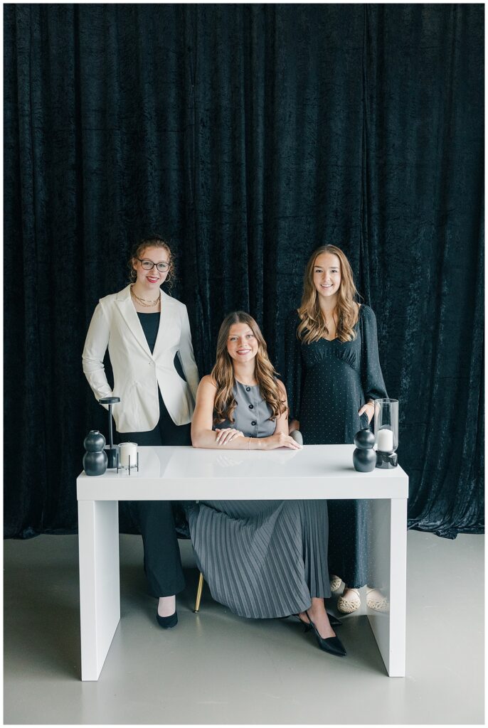 Branding photo of a business team of three women at a white table in front of a black curtain backdrop, portraying a professional and modern workspace.