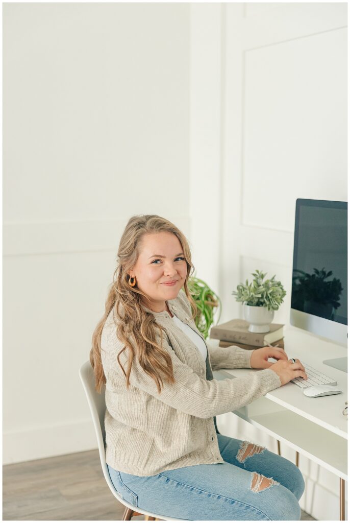 Bright office brand photo of a smiling woman working at her desktop computer, styled with greenery and neutral tones.