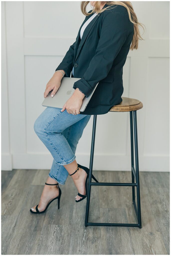 Close-up brand photo of a woman in jeans and heels holding a laptop while sitting on a modern wooden stool in a white office space.