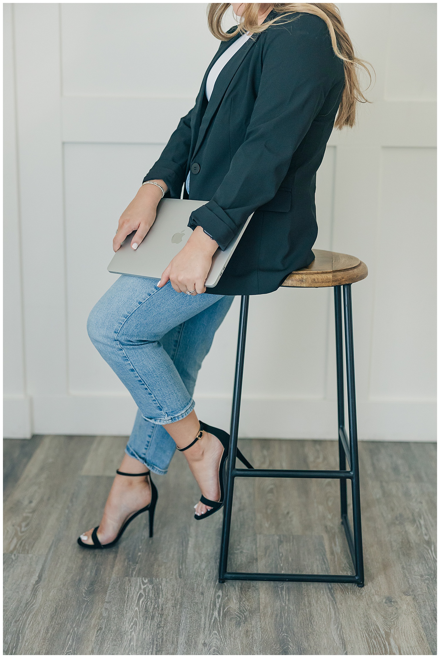 Close-up brand photo of a woman in jeans and heels holding a laptop while sitting on a modern wooden stool in a white office space.
