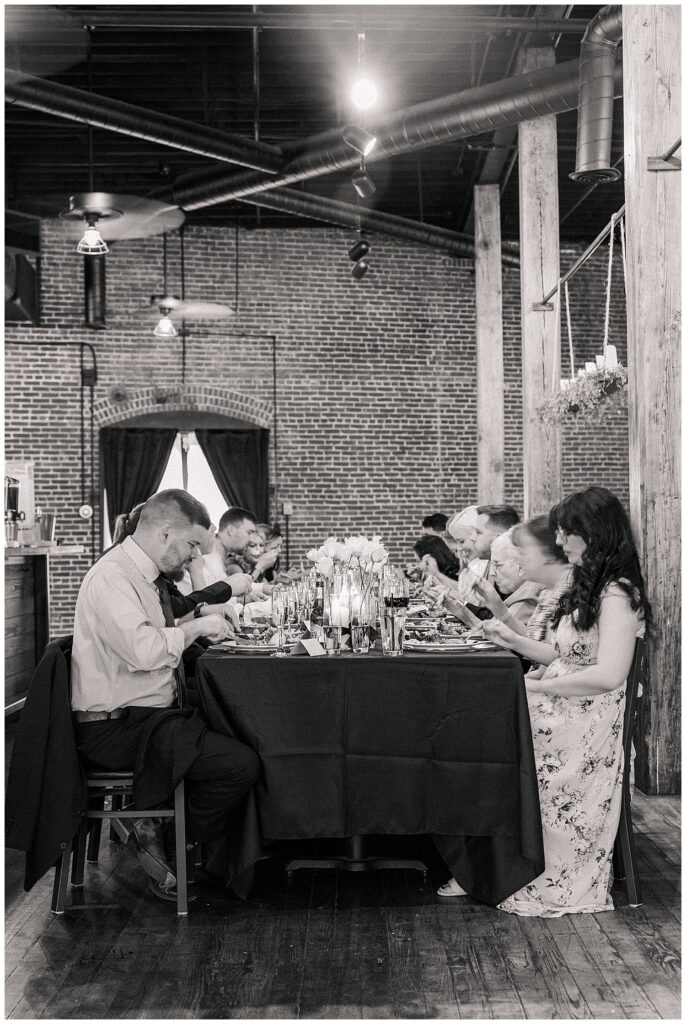 Black-and-white image of wedding guests dining at an intimate reception inside an industrial-style brick venue.