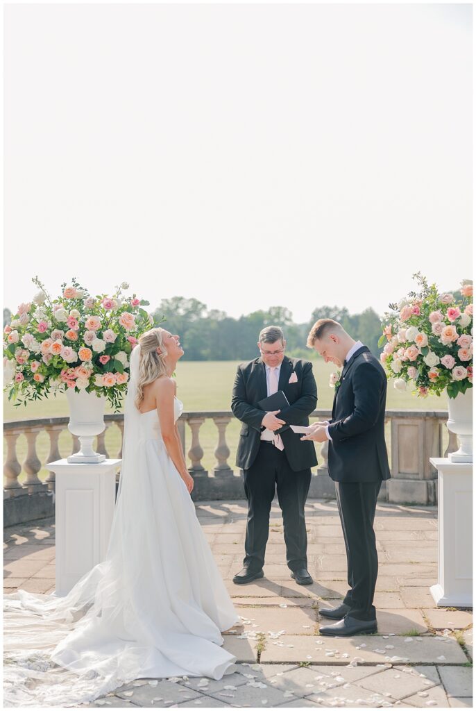 Bride and groom reading their vows on a sunlit terrace, framed by large blush floral arrangements.