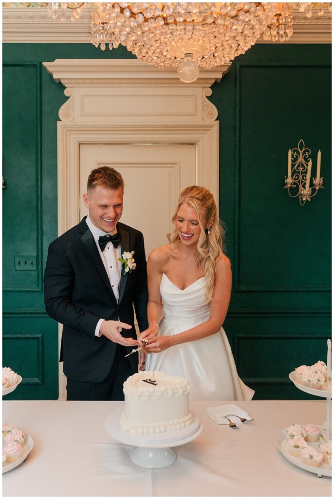 Bride and groom cutting their wedding cake in front of a green wall with elegant decor.