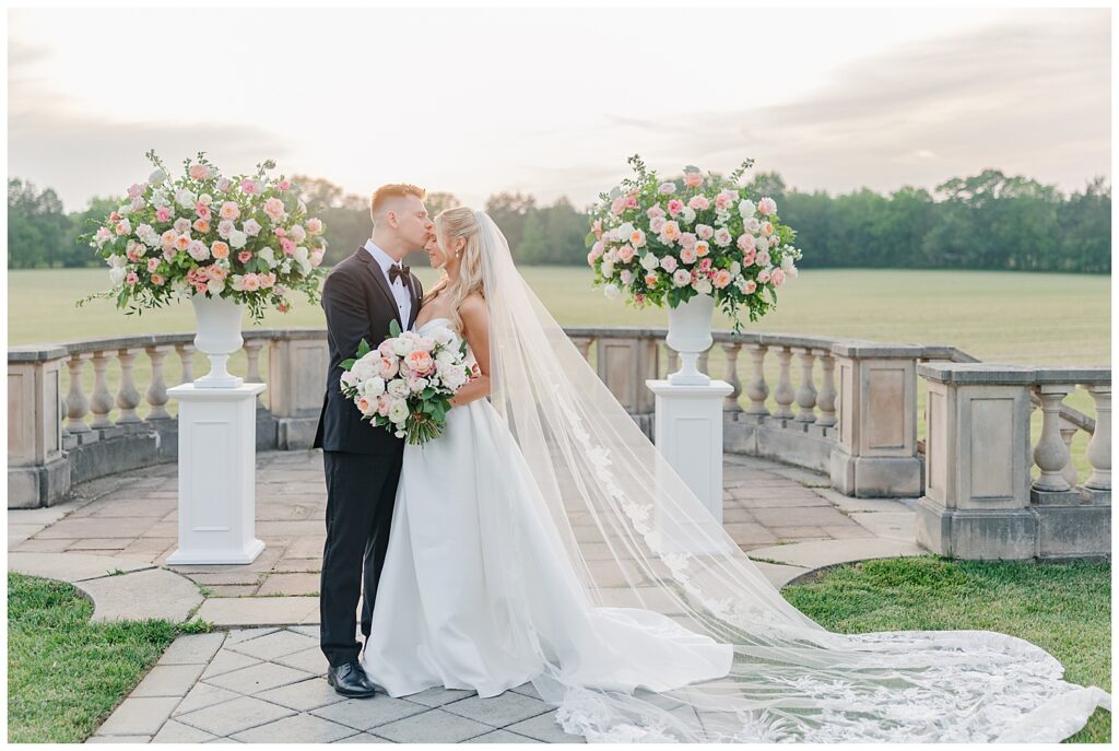 Romantic portrait of bride and groom embracing on a stone terrace surrounded by floral arrangements at sunset.