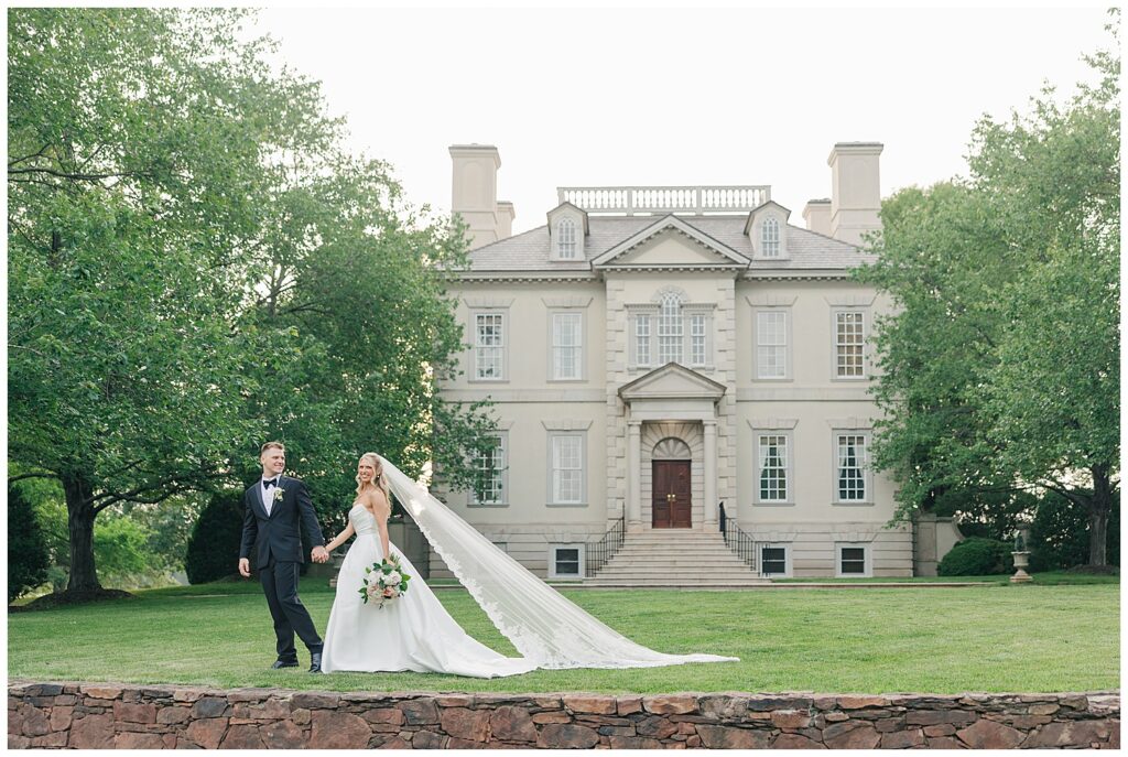 Bride and groom walking hand-in-hand in front of a historic mansion with the bride’s long veil trailing behind.