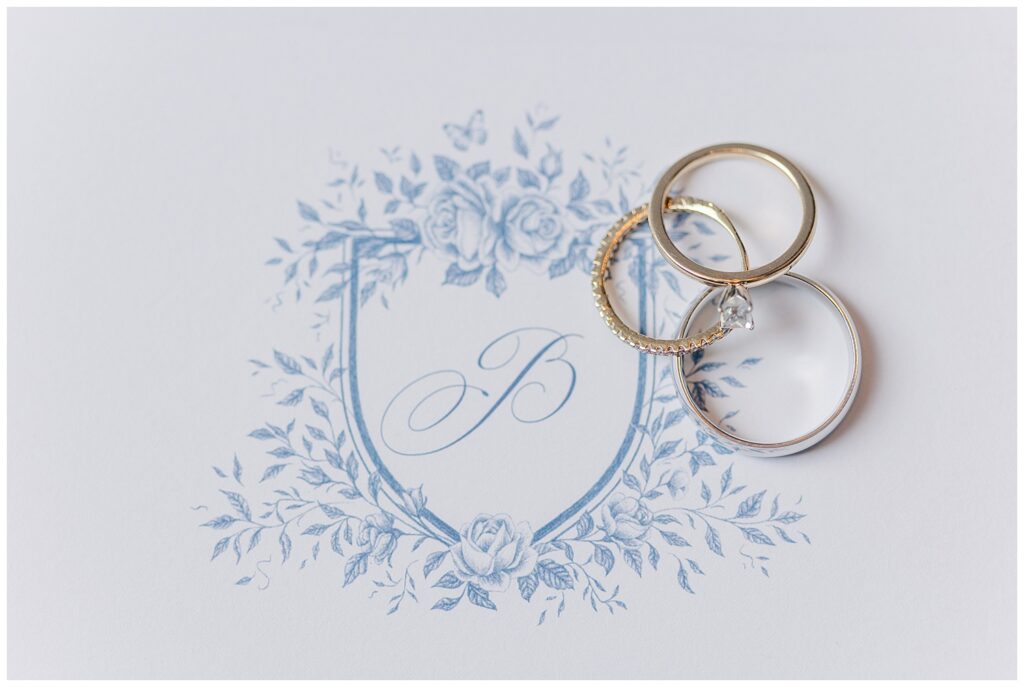 Flat lay of wedding rings placed on a custom blue wedding crest featuring the couple’s initial.