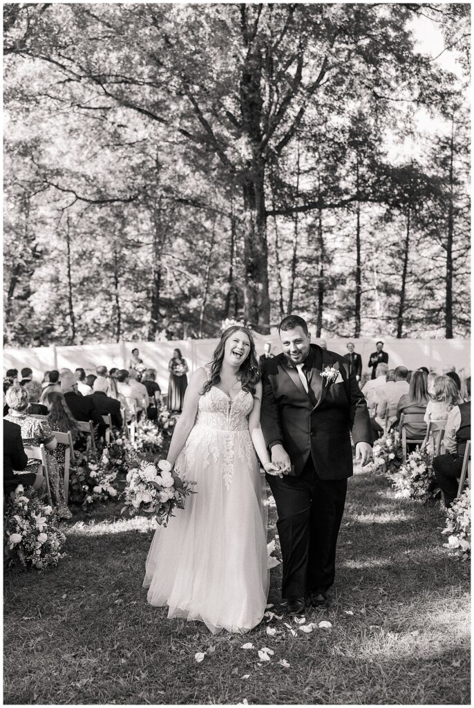 Bride and groom walking up the outdoor ceremony aisle at Ellis House surrounded by trees and guests.