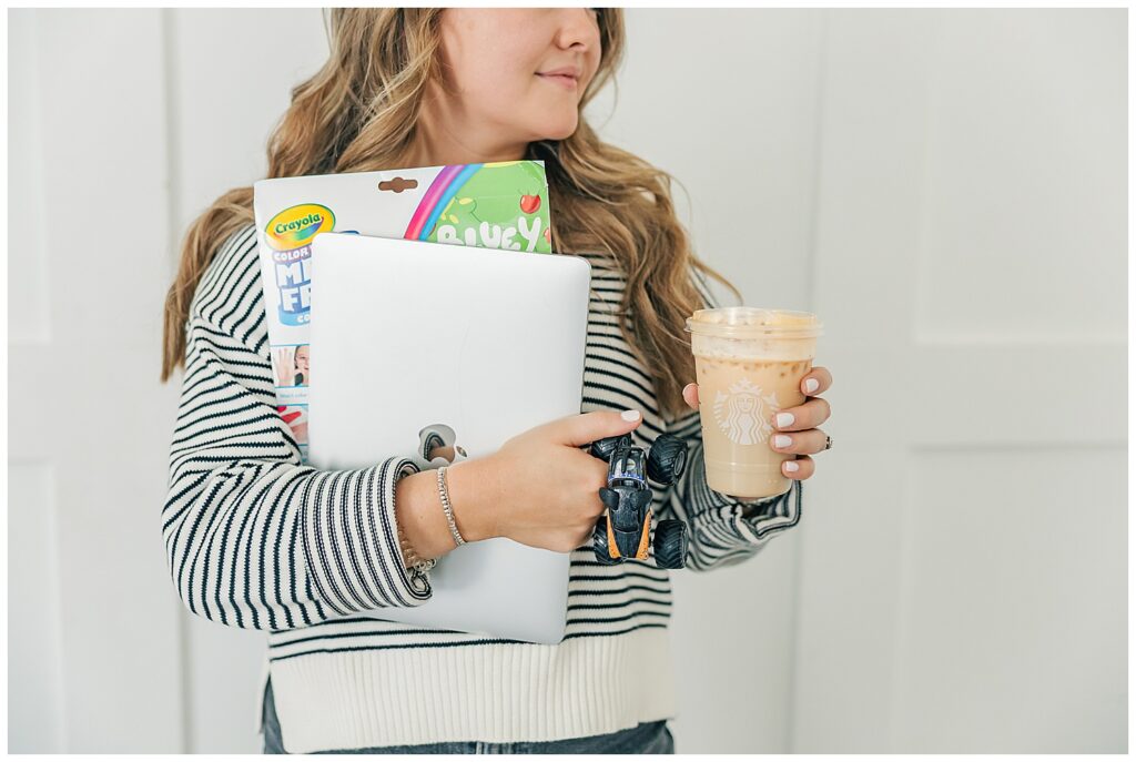 Close-up of Liz holding her laptop, client folders, coffee, and toddler toys—emphasizing motherhood and multitasking in business.
