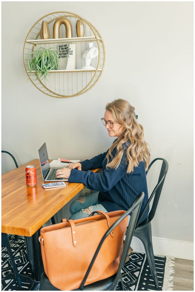 Virtual assistant Liz Agnellini types on her laptop at a warm wood table with plants and natural light. Intentional workspace for her remote business.