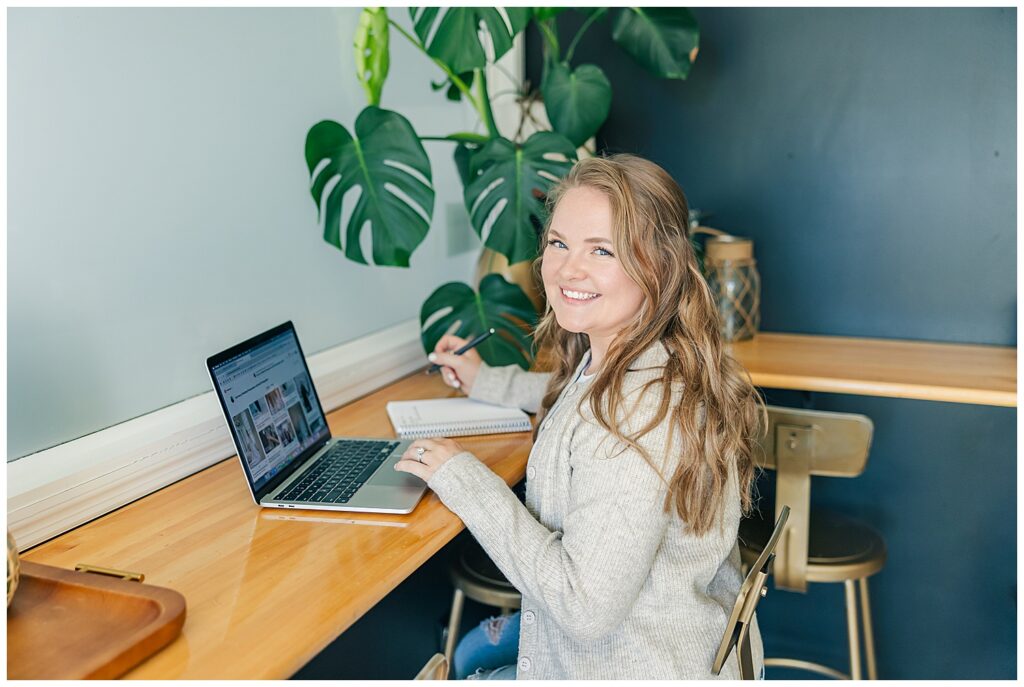 Liz Agnellini smiles at her laptop during a brand session in a cozy coffee shop setting, showcasing her virtual assistant work-from-anywhere lifestyle.